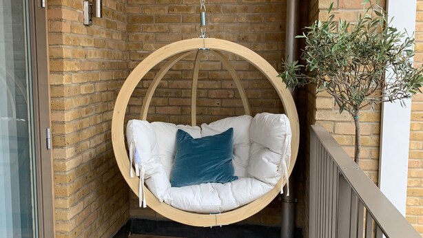 Una poltrona da esterno in legno con cuscini bianchi e un cuscino color petrolio, appesa tramite una catena al soffitto, con una parete in mattoni sullo sfondo.