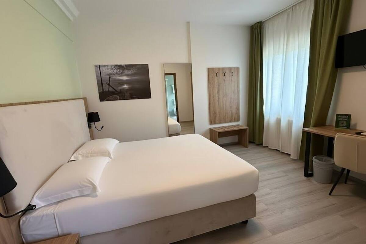 A modern hotel room with a large bed, a desk, a chair, a TV, and a painting on the wall.