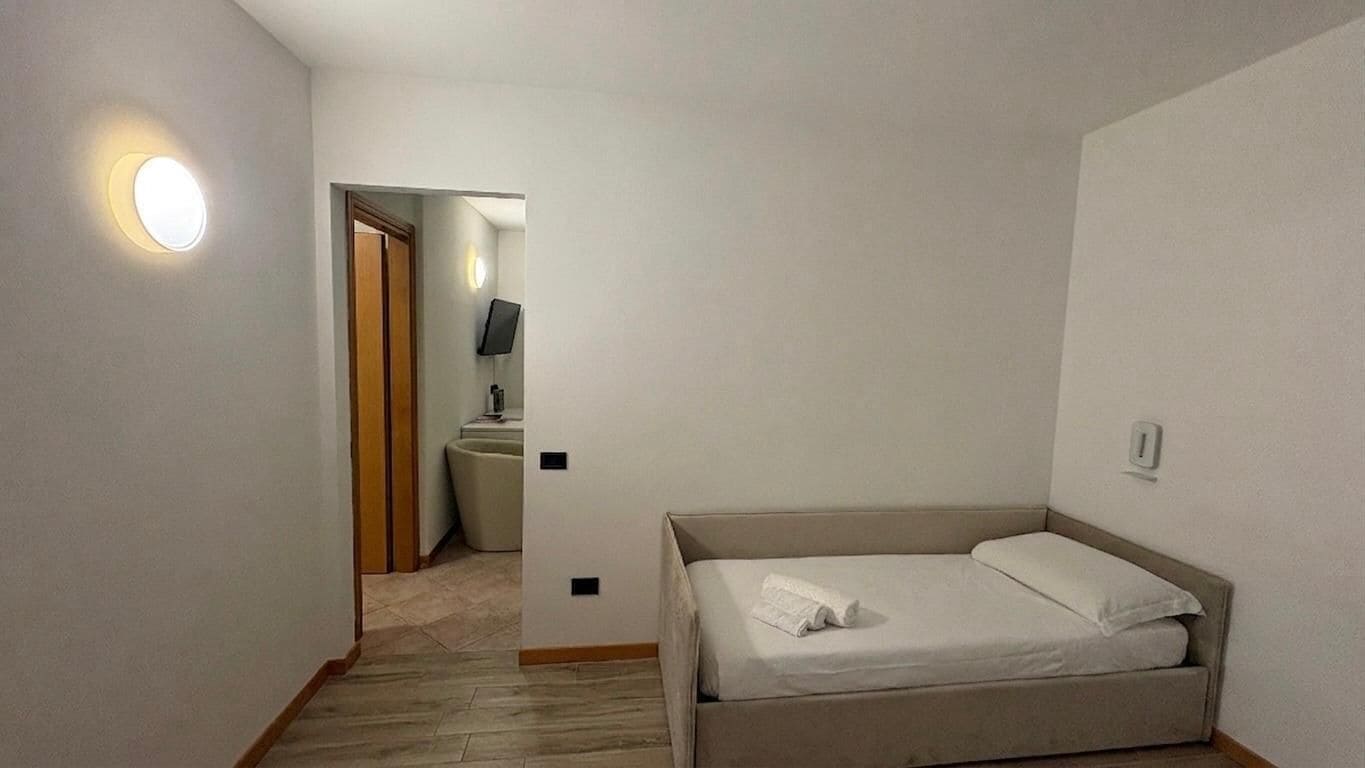 Une petite pièce propre, avec un lit, une lampe fixée au mur et une porte donnant sur une salle de bain.