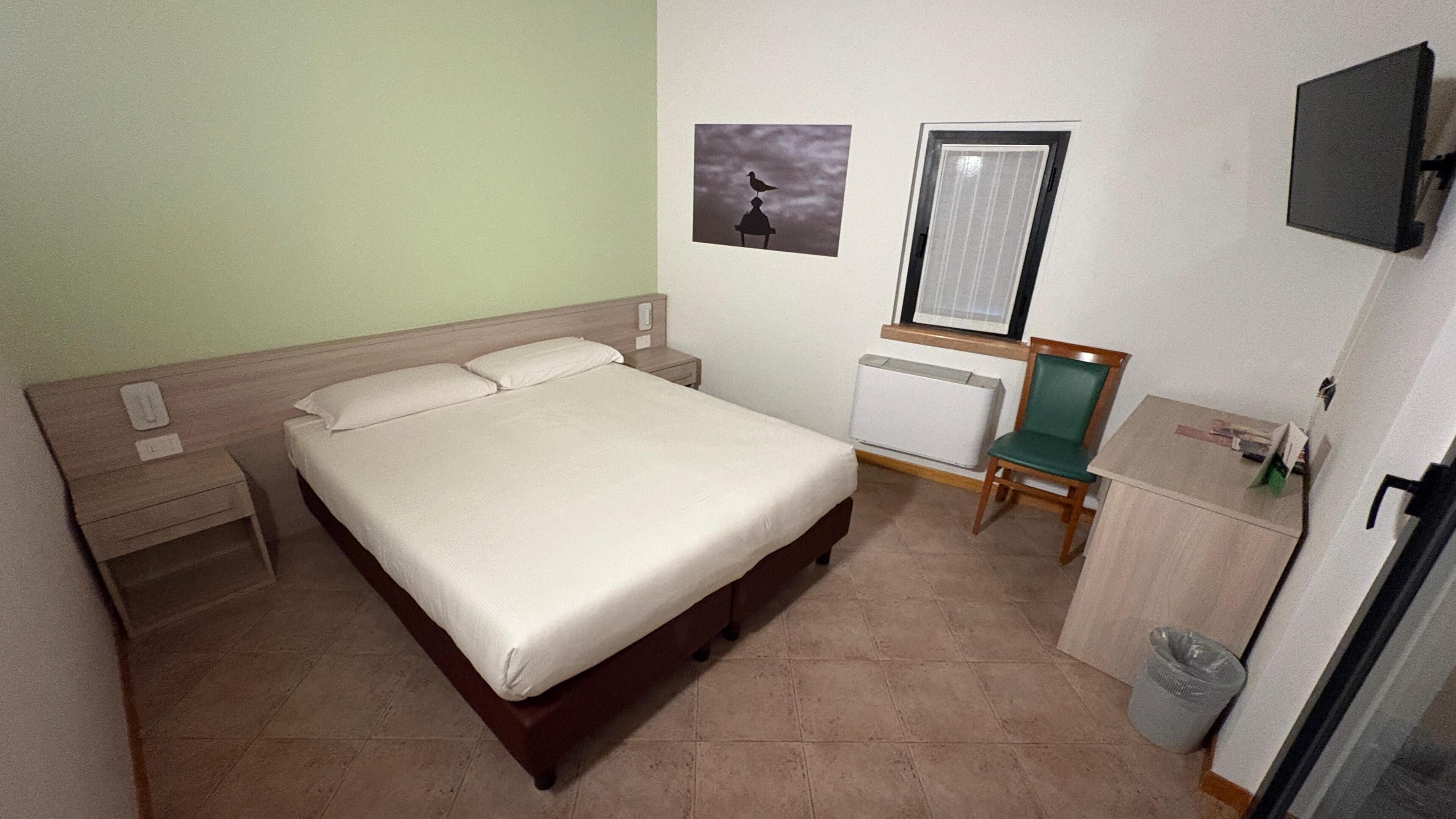 Quarto de hotel com cama, poltrona, escrivaninha, radiador e um quadro na parede.