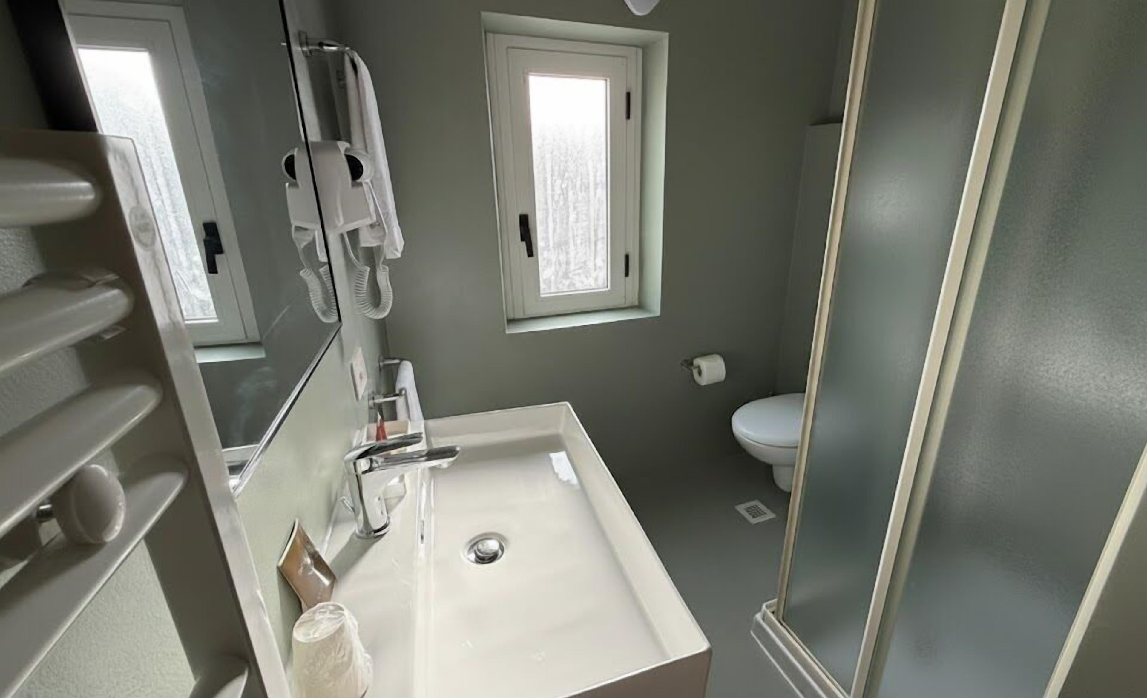 A bathroom with a white sink, a toilet, and a shower. There is a window above the toilet and a towel rack above the sink.