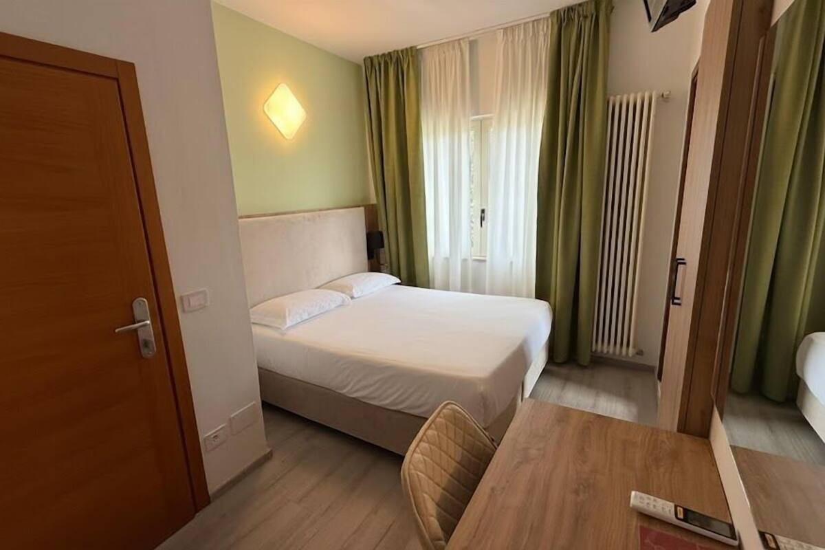 A hotel room with a bed, a desk, a chair, a nightstand, and a window with curtains.