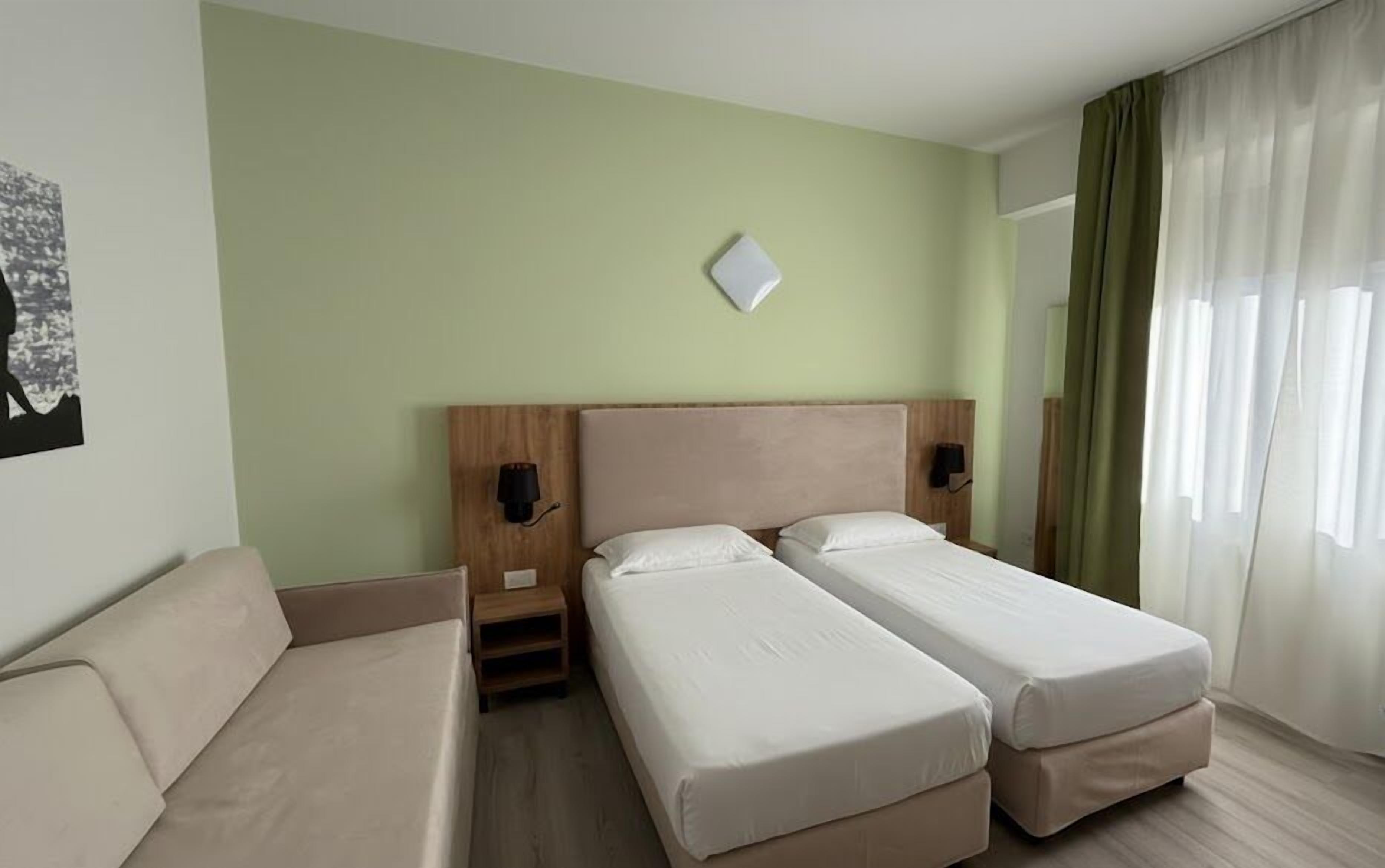 A hotel room with two single beds, a wooden headboard, a small bedside table, a wall-mounted light, and a window with curtains.