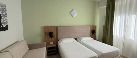 A hotel room with two single beds, a wooden headboard, a small bedside table, a wall-mounted light, and a window with curtains.