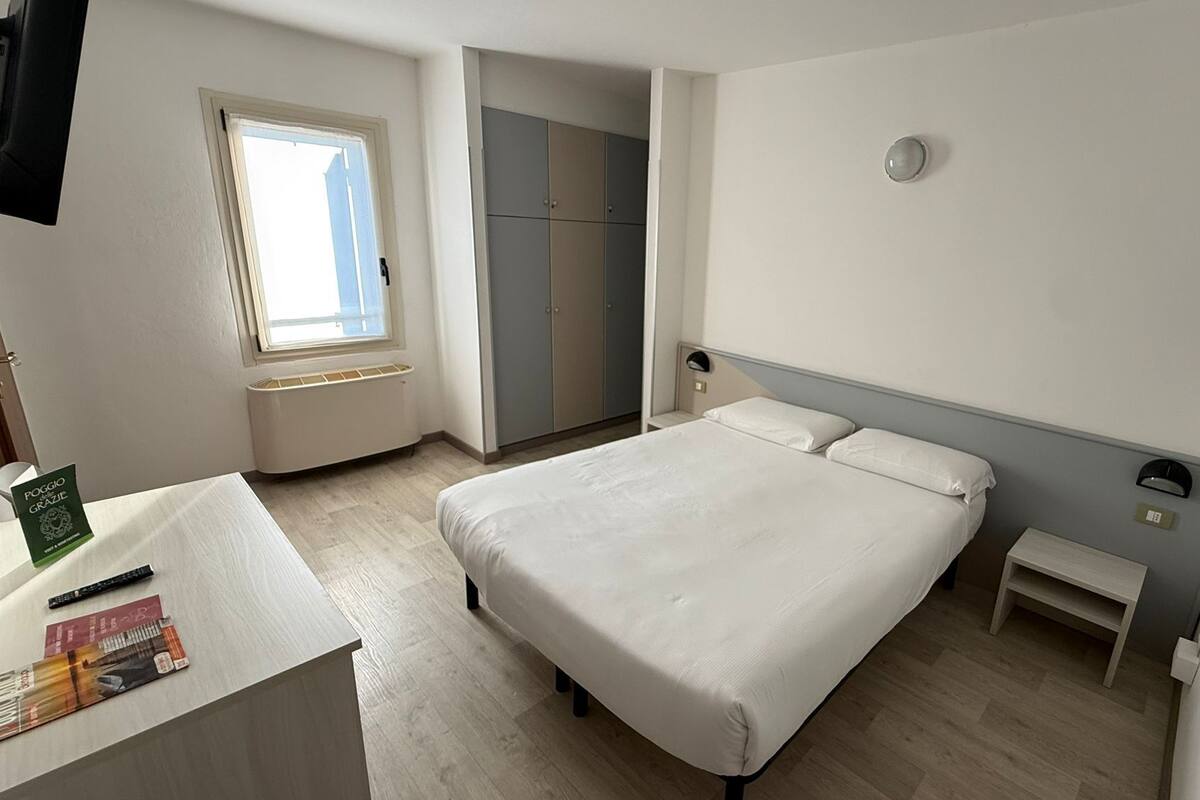 A hotel room with a bed, bedside tables, a desk with a chair, a window, a wardrobe, and a television.