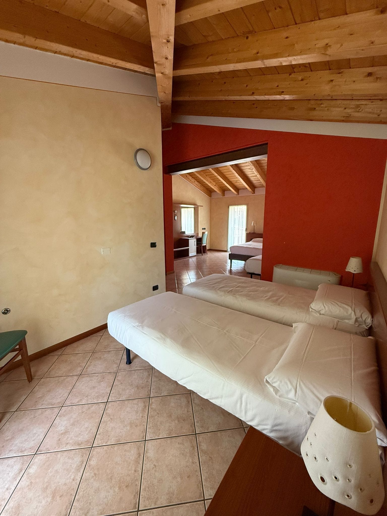 A room with two beds, a small table, and a chair. There is a wooden ceiling and a red wall.