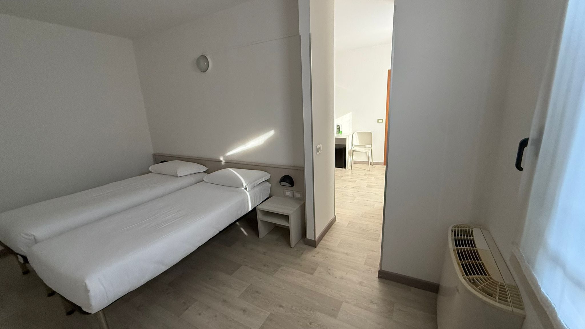 A small, single-bed room with a wooden floor, a bedside table, and a chair in the background.