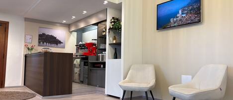 Un hall d’hôtel comprenant un comptoir de réception, deux fauteuils, un appareil à café, un téléviseur fixé au mur et un tableau encadré.