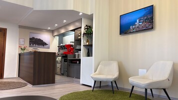 Un vestíbulo de hotel con mostrador de recepción, dos sillones, una máquina de café, un televisor montado en la pared y un cuadro enmarcado.