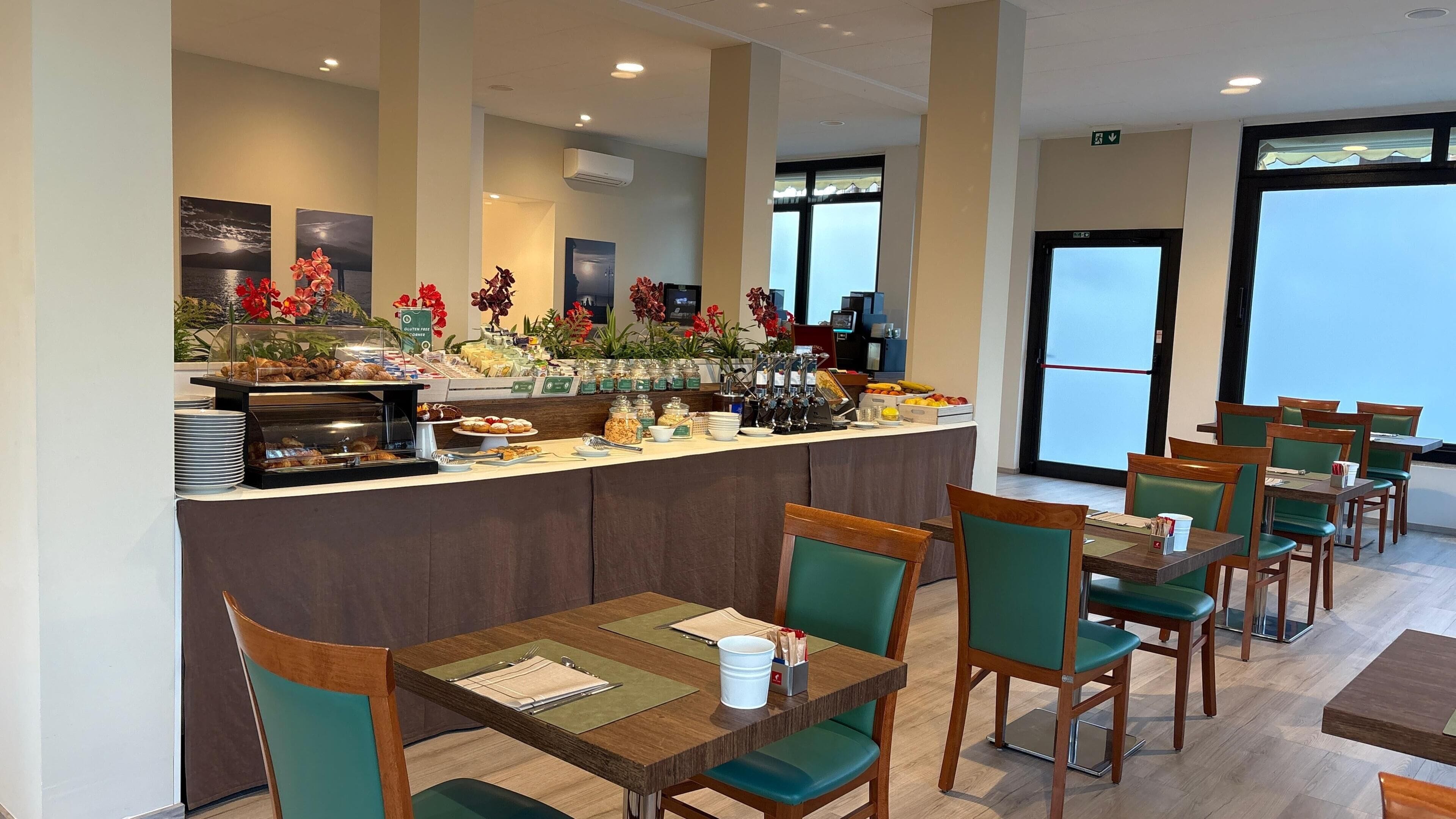 Espaço de refeições de hotel com buffet, cadeiras verdes e vista pela janela.