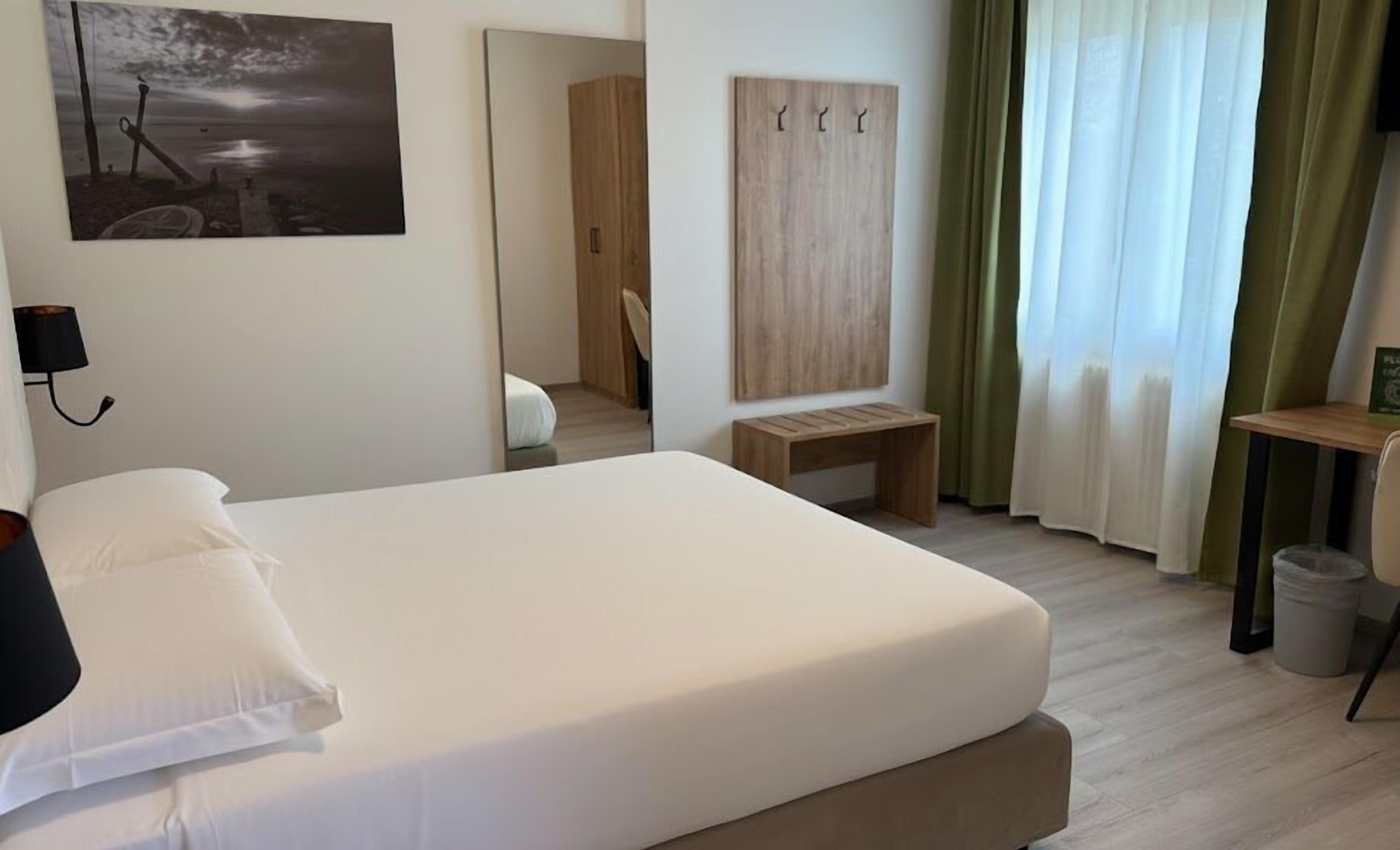 A hotel room with a bed, a desk, a chair, a wardrobe, and a painting on the wall.