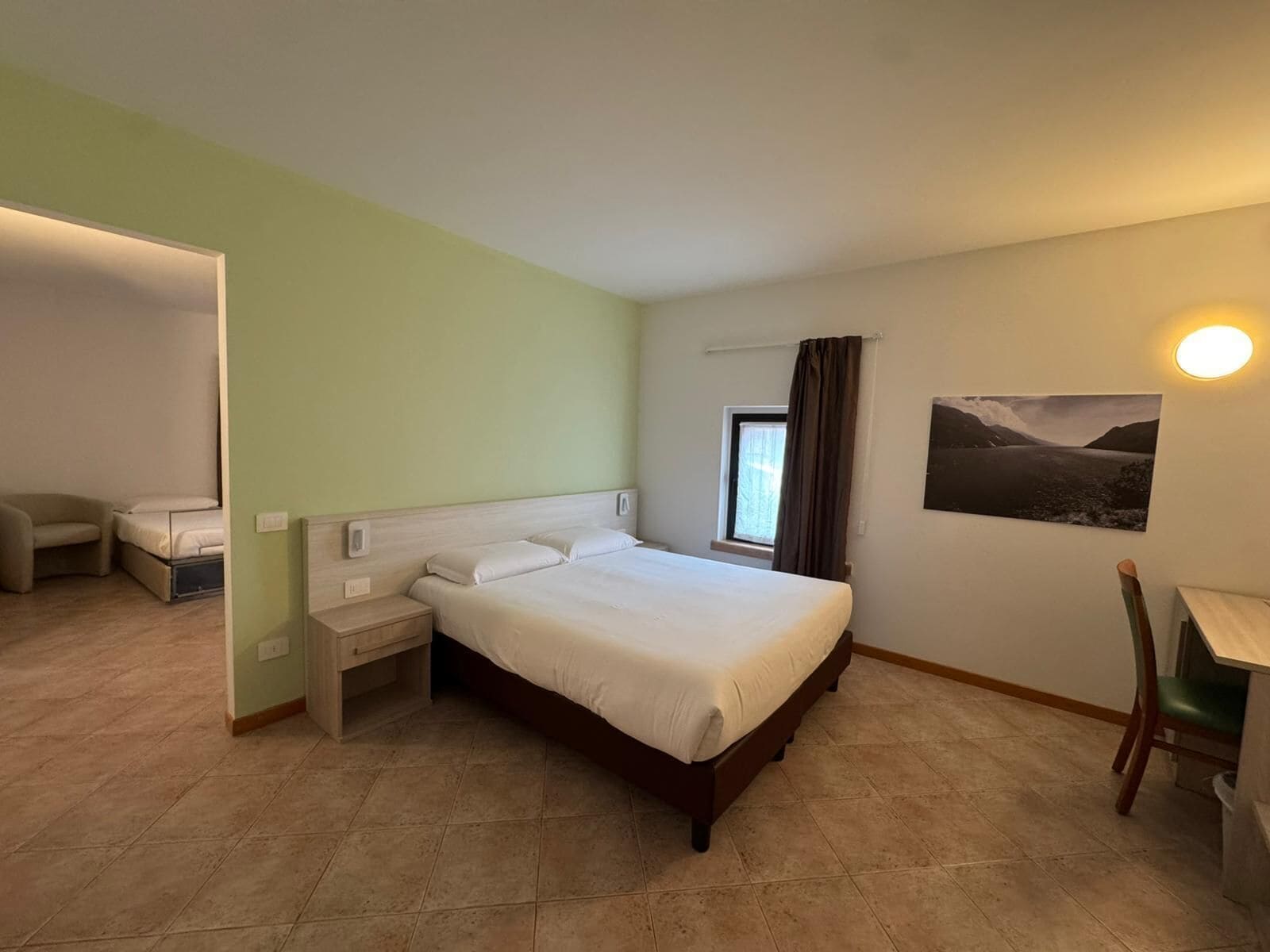 Ett hotellrum med en säng, ett skrivbord, en stol, ett fönster och en tavla på väggen.