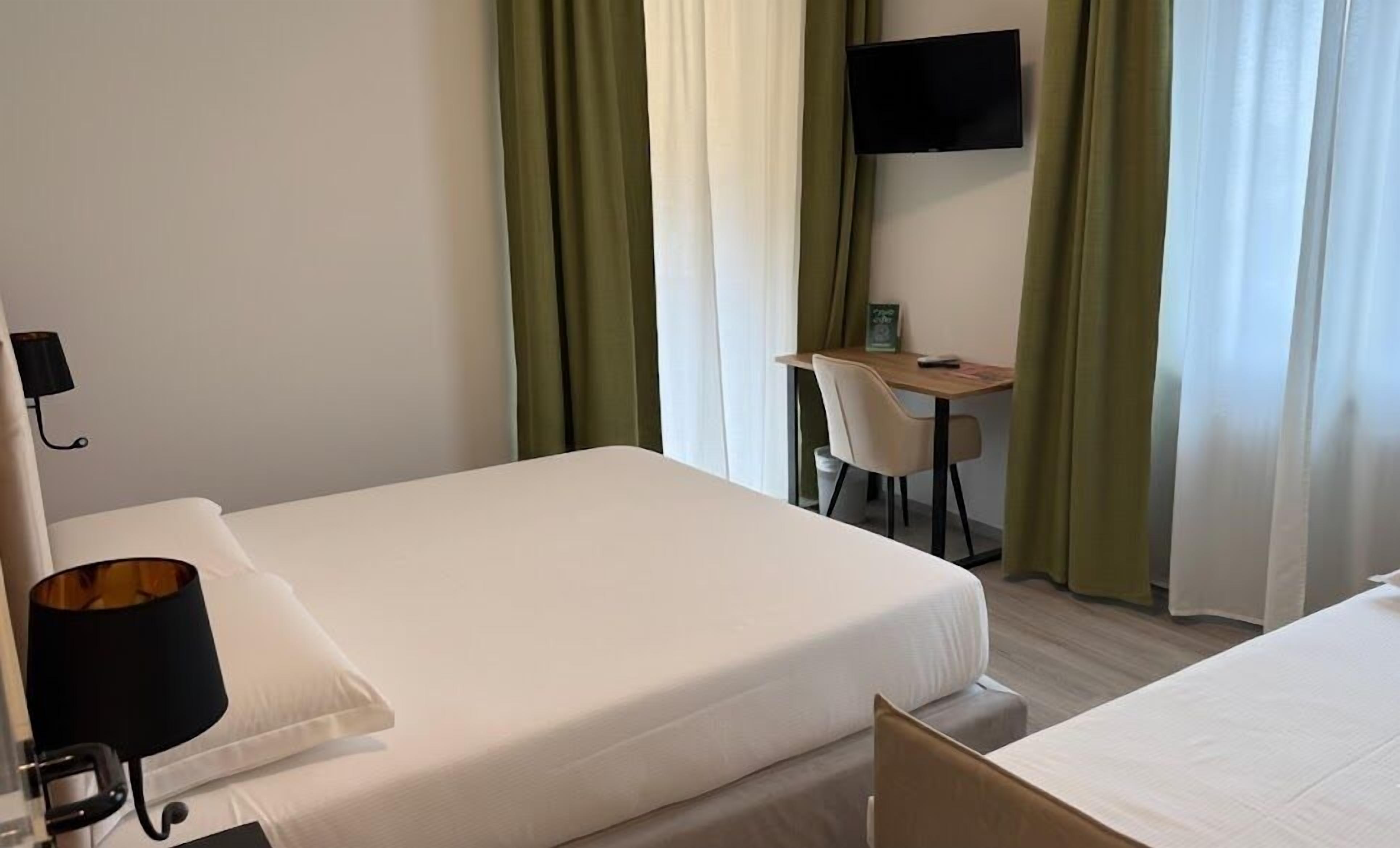 Habitación de hotel con cama, escritorio y silla, televisor en la pared y cortinas verdes.