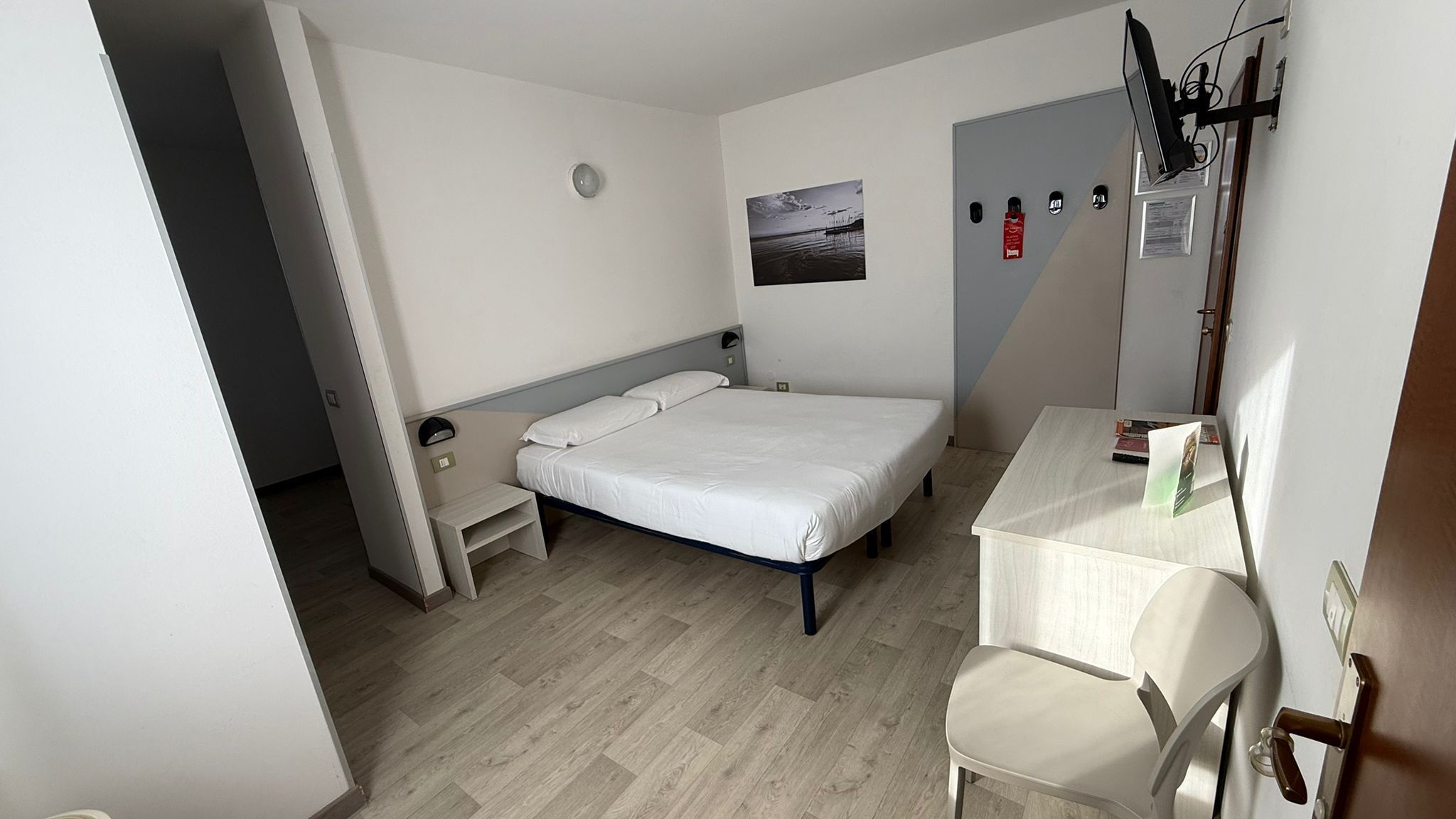 A compact hotel room with a single bed, a small desk, and a chair. There is a window with a view of a beach, a fire extinguisher, and a door with a handle.