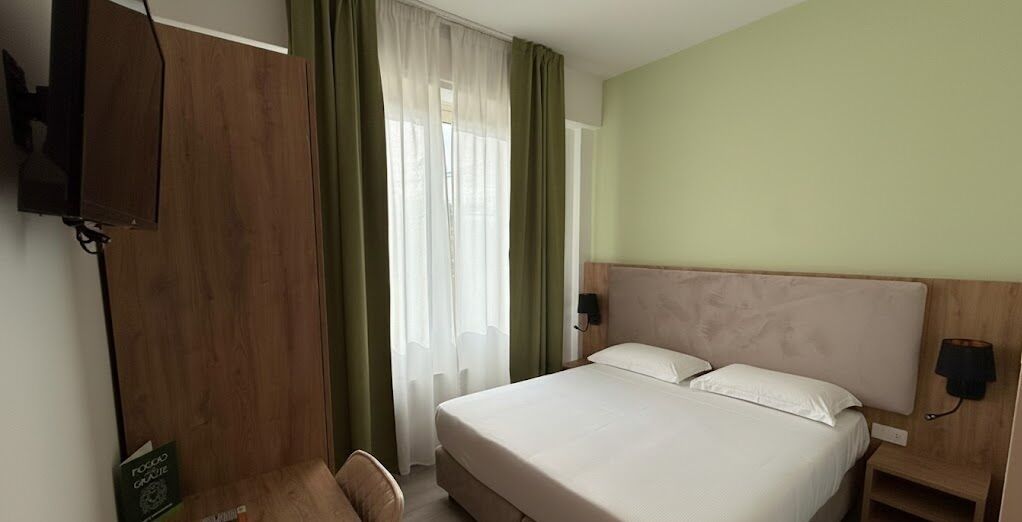 Habitación de hotel con cama, escritorio, silla, televisor y ventana con cortinas.