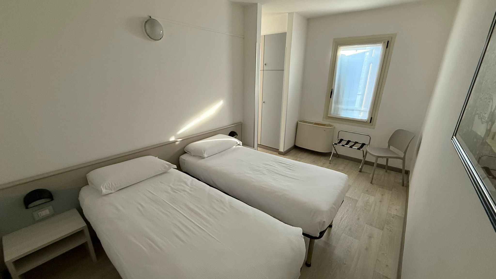 Una habitación pequeña y limpia con dos camas individuales, una silla, un escritorio y una ventana.