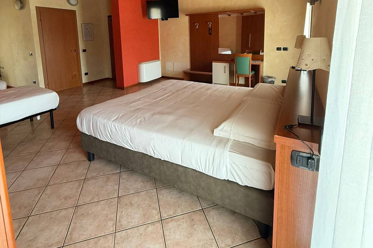 A bedroom with a large bed, a desk, a chair, a TV, and a bathroom visible in the background.