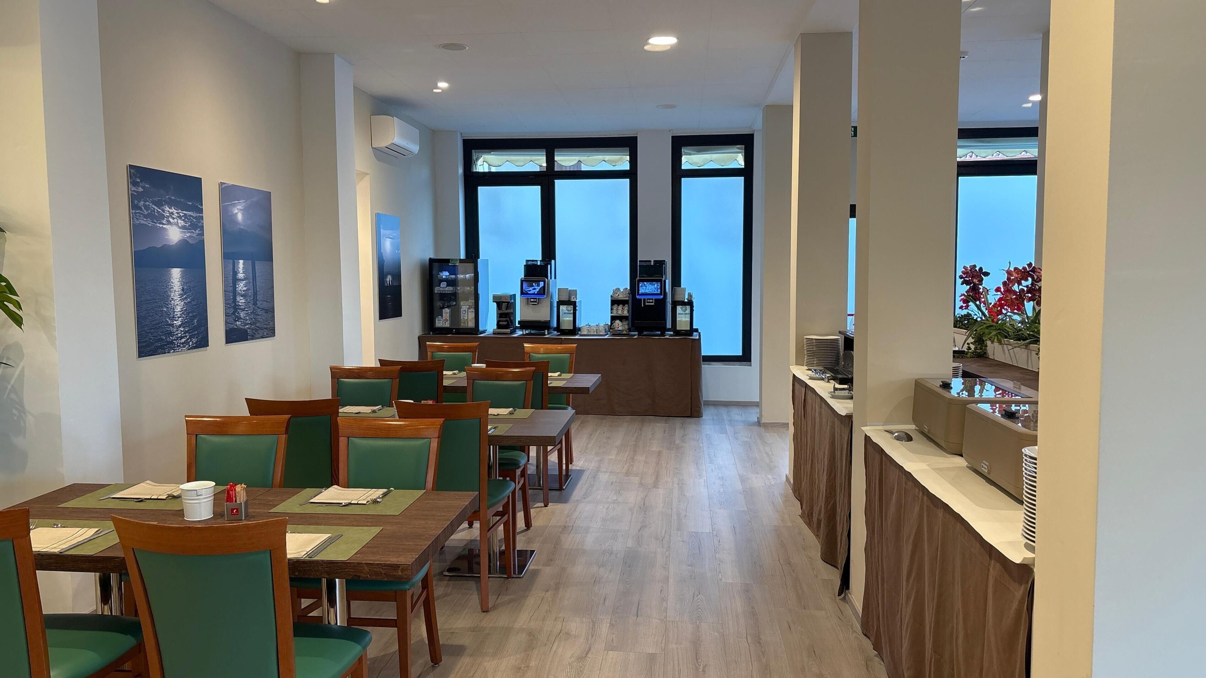 A hotel dining area with wooden tables and green chairs, a bar with a counter, and large windows with a view.