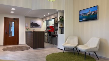 Zona de recepción de hotel con puerta de madera, mostrador de recepción, dos sillas blancas, alfombra verde, máquina de café y televisor mostrando una vista panorámica.