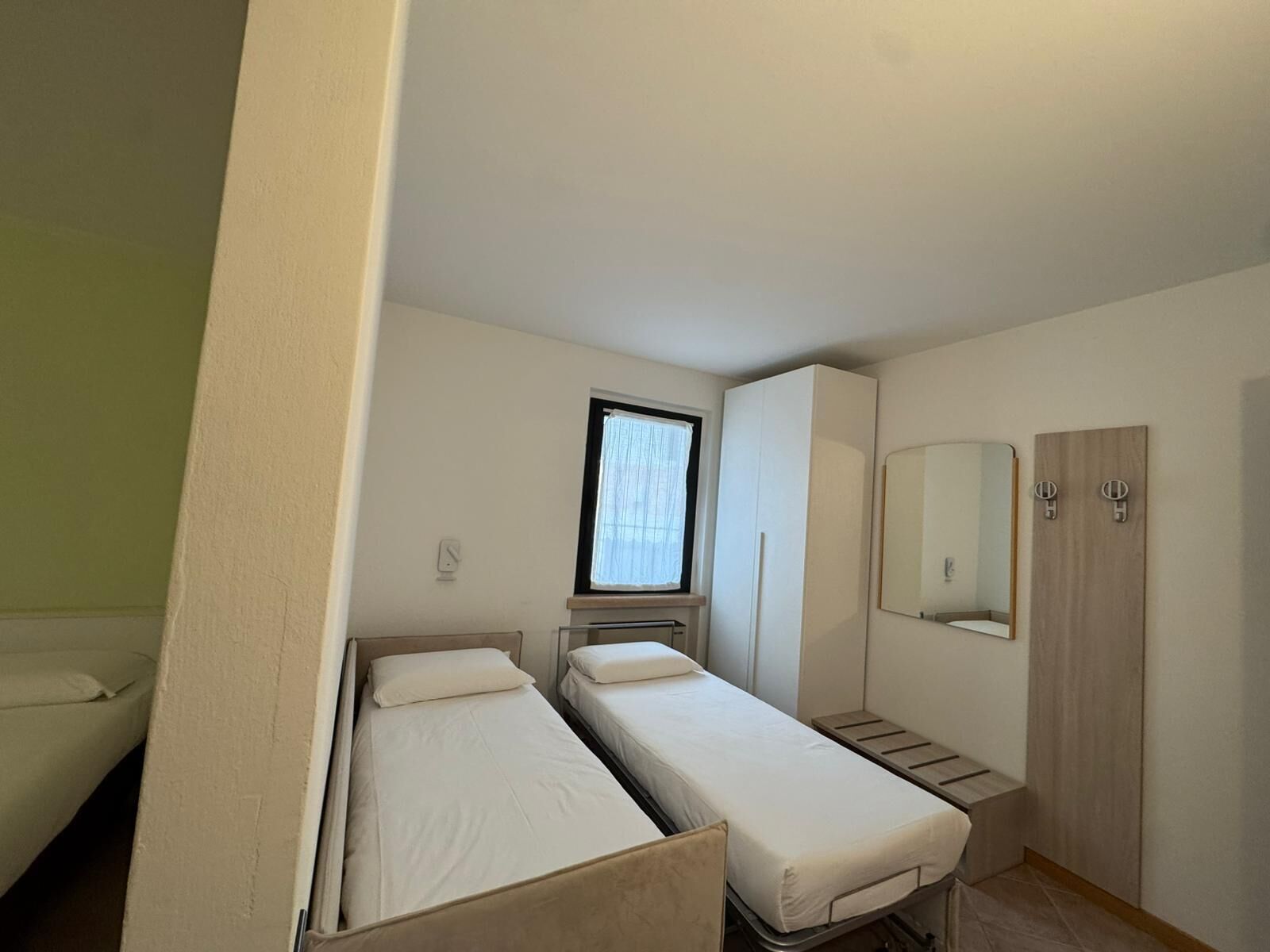 Una habitación compacta con dos camas individuales, una ventana pequeña con cortinas, un espejo y un armario.