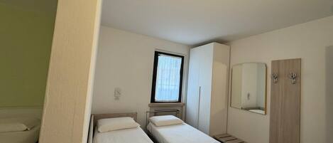 Una habitación compacta con dos camas individuales, una ventana pequeña con cortinas, un espejo y un armario.