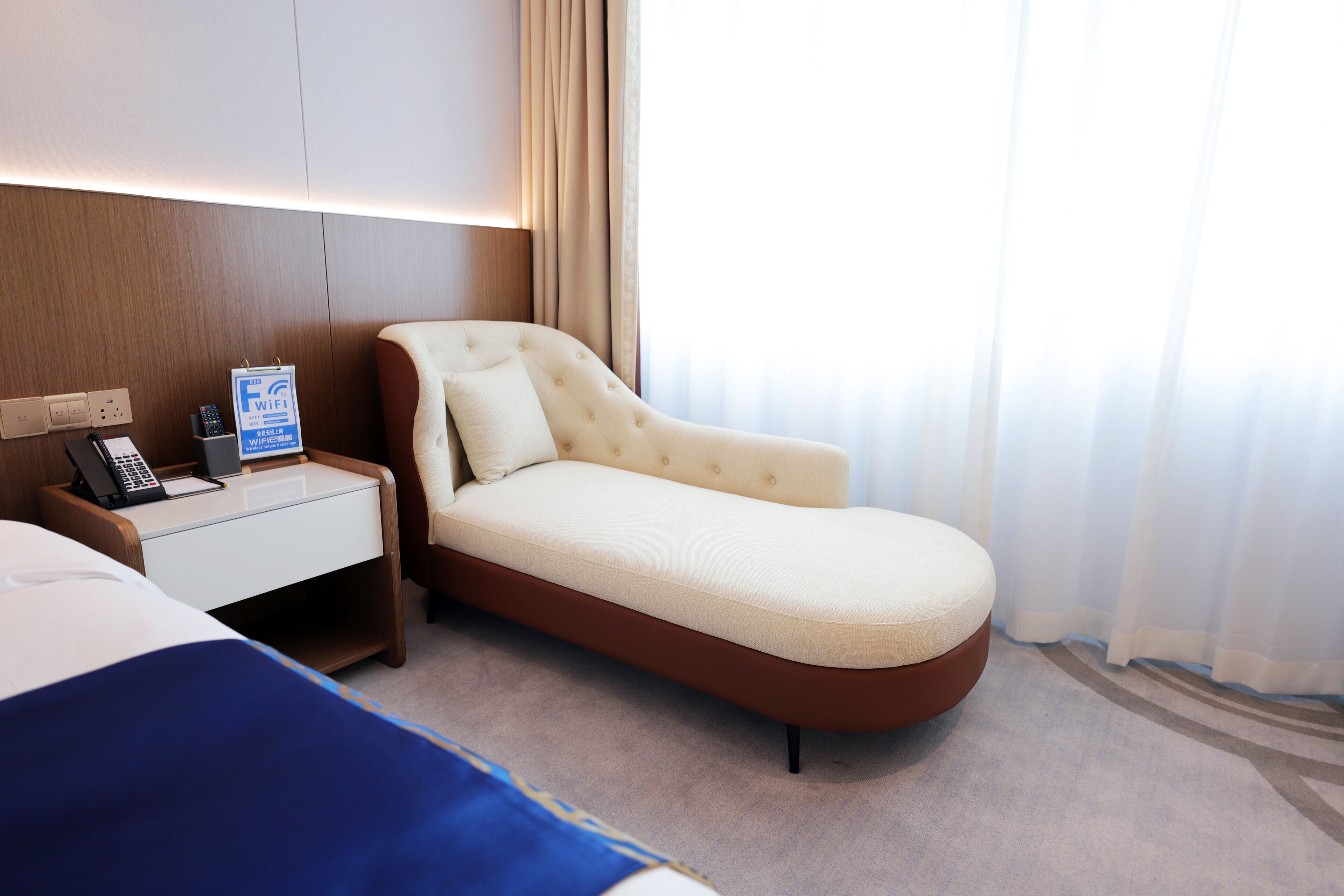 A hotel room with a single bed, a desk with a phone and a sign indicating WiFi availability, and a window with sheer curtains.