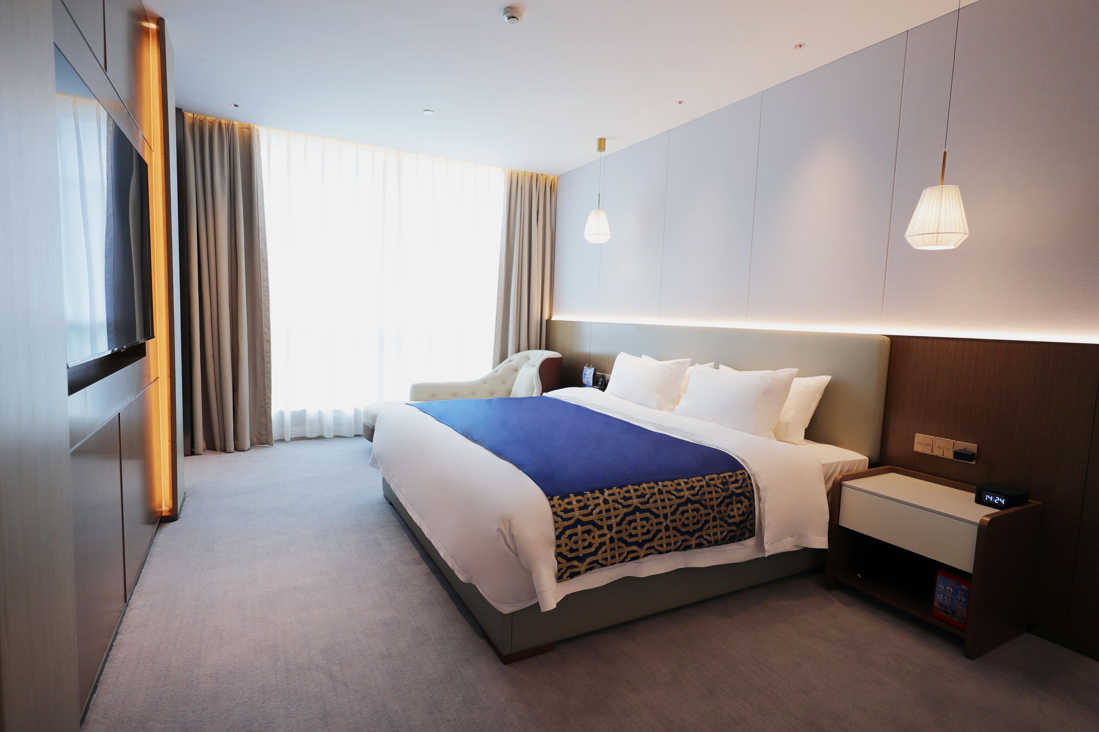 Una habitación de hotel moderna con una cama grande, una mesita de noche con un reloj, un televisor montado en la pared y un amplio ventanal con cortinas.
