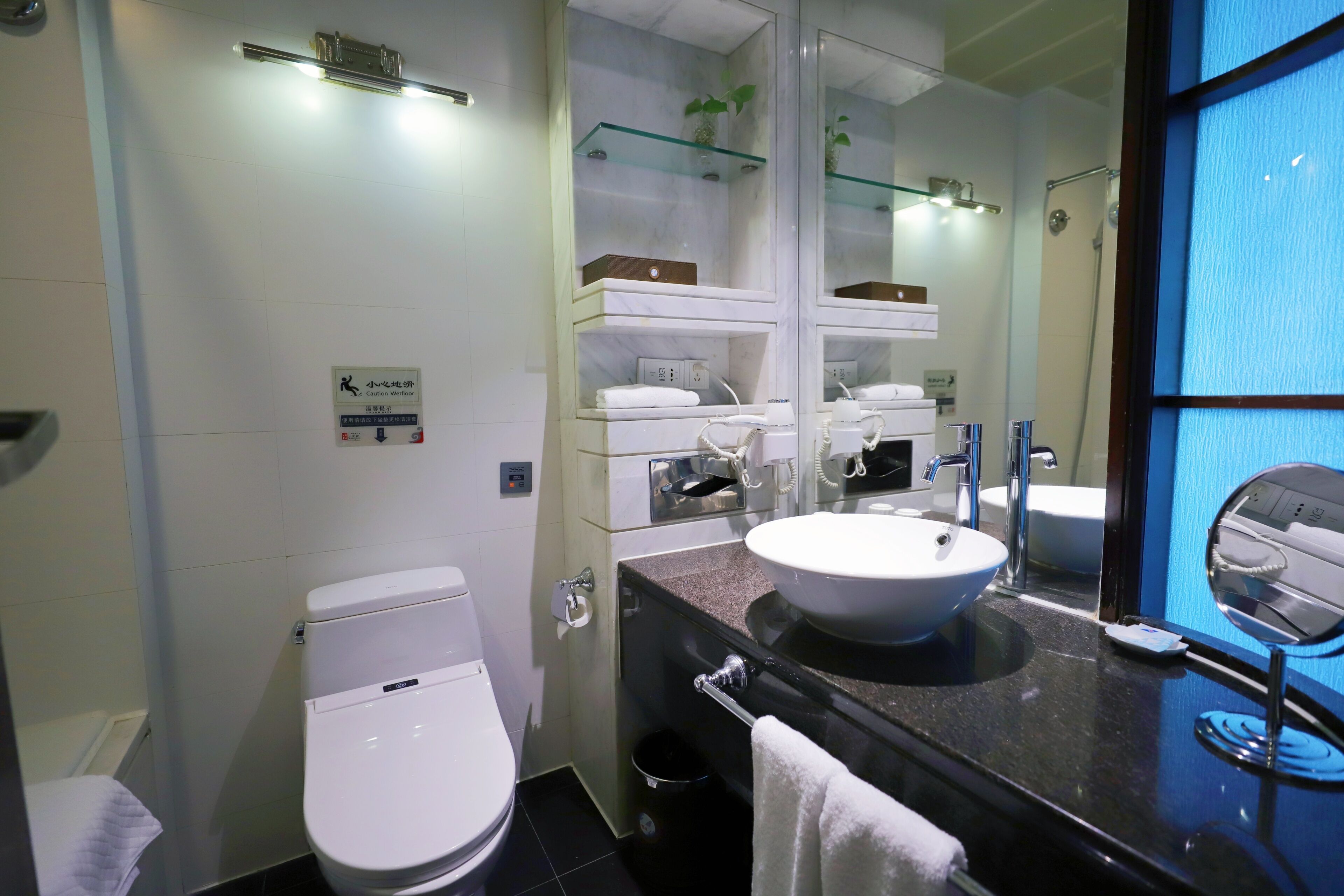 Un baño moderno con inodoro, lavabo, espejo y repisa de vidrio con artículos de aseo.
