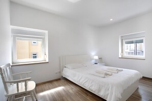 Une chambre à coucher avec un lit, une chaise, deux fenêtres et une table de chevet avec une lampe.