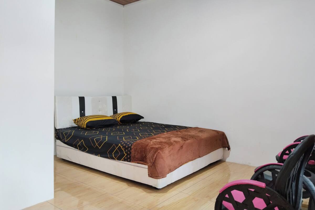 A small, neatly arranged bedroom with a single bed, a brown blanket, and a chair with a patterned seat.