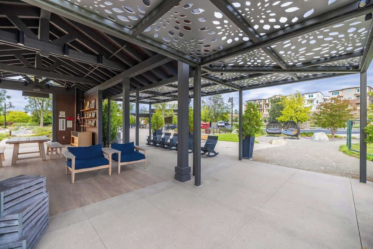En utomhusplats med sittmöbler, en pergola som ger skydd, träbord, blå sittdynor på stolarna och en hög med tidningar.