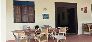 A dining area with a wooden table and four wicker chairs, a window with a woven pattern, and a door leading to another room. - Furaha Beach Bungalows by Serenity (Jambiani)