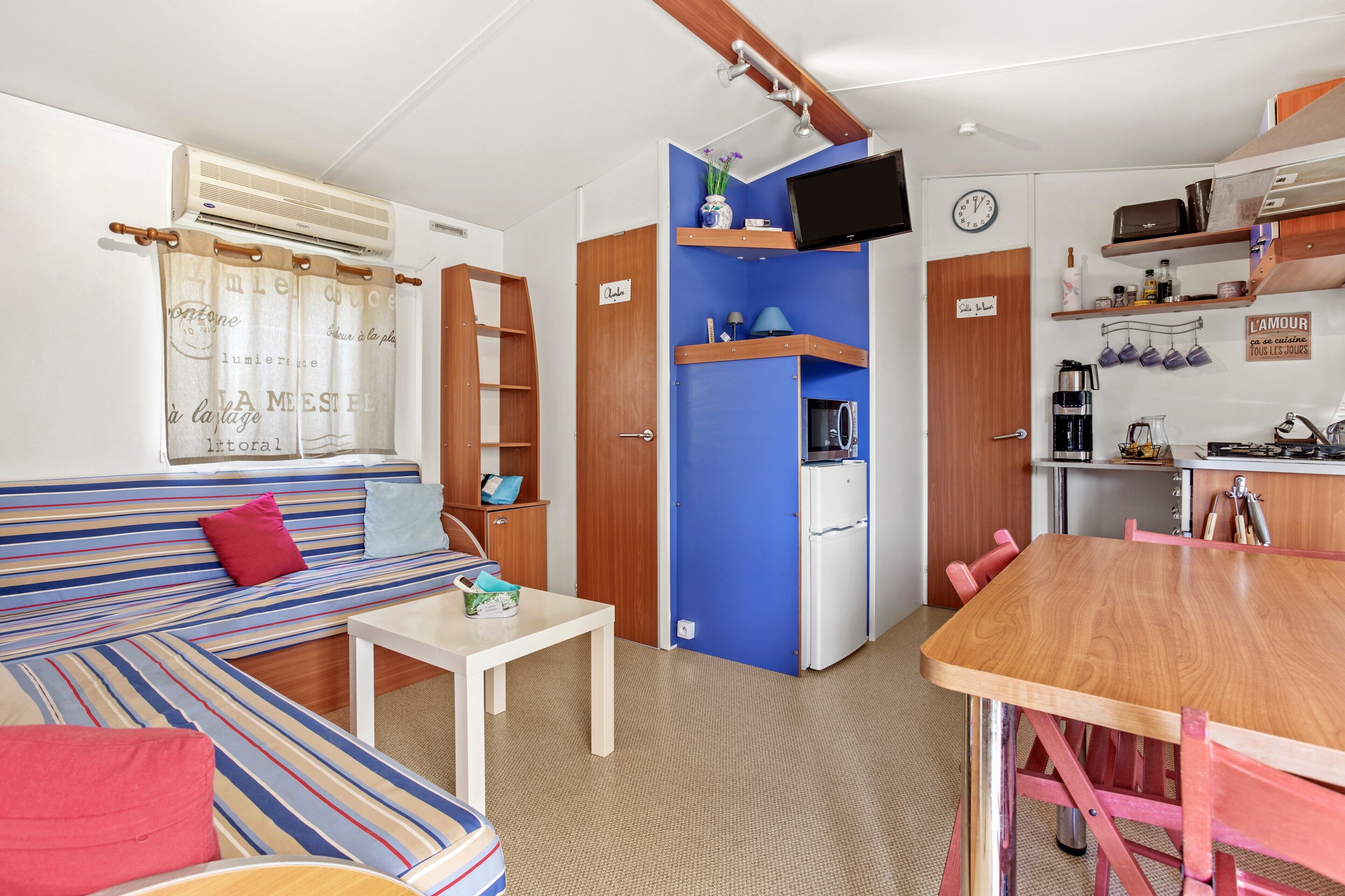 A compact room with a sofa, a small table, a kitchenette, and a TV mounted on the wall.