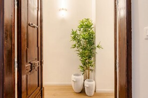 A hallway with a wooden door, a wall-mounted light fixture, a bamboo plant, and two white ceramic pots.