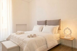 A neatly made bed with white bedding and two rolled towels, a bedside table with a lighted decorative object, and a bench at the foot of the bed. - Simona - Italian Living (Monopoli)