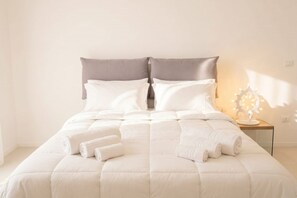 A neatly made bed with white bedding and pillows, a bedside table with a lamp, and a wall-mounted light fixture.
