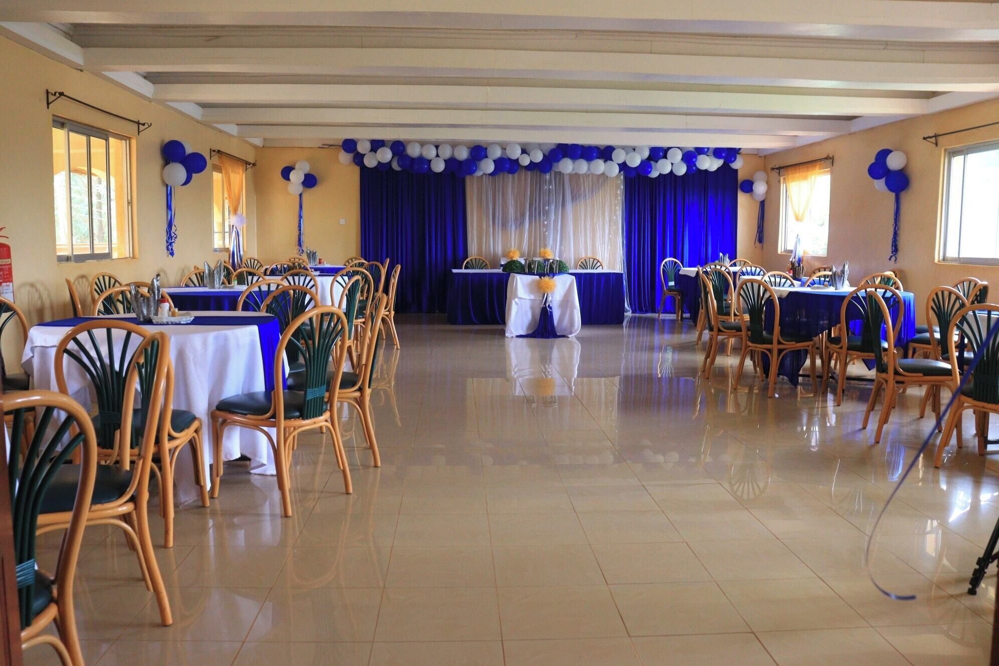 A banquet hall with tables set for dining, wooden chairs, and a stage with a blue curtain.
