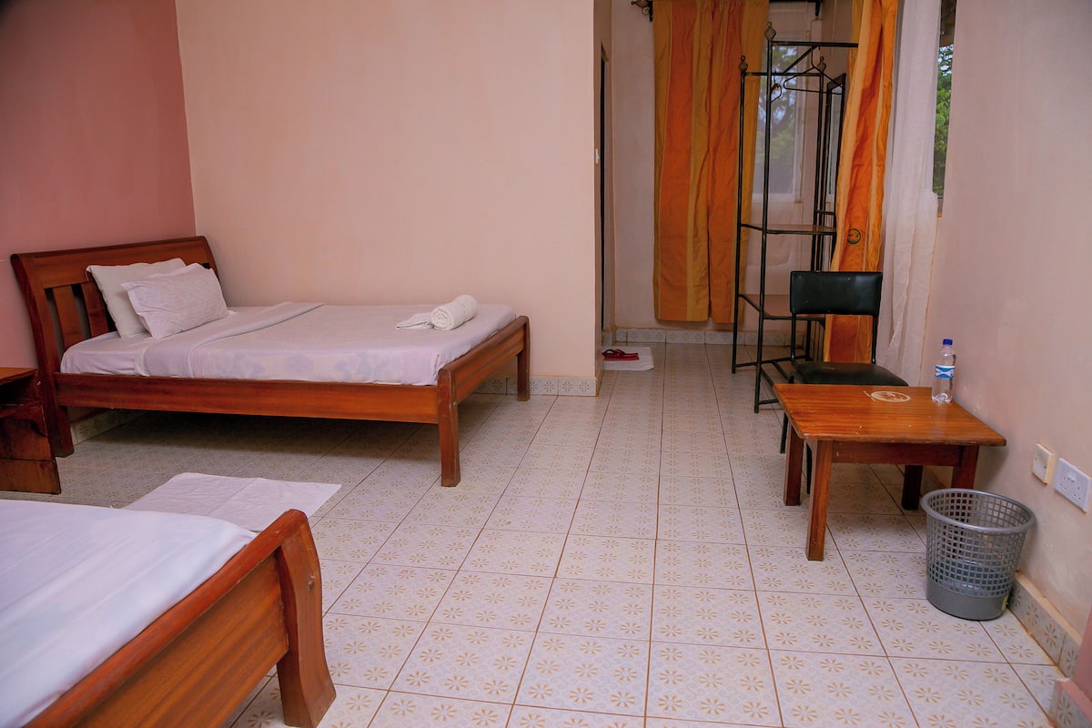 A bedroom with two beds, a wooden table, a chair, and a trash bin.