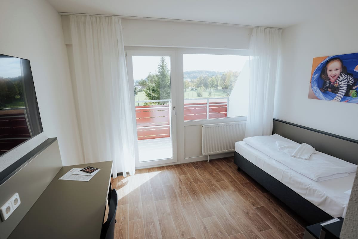 Una habitación de hotel con una cama, un escritorio con computadora, una ventana con cortinas y vista a un edificio rojo en el exterior.