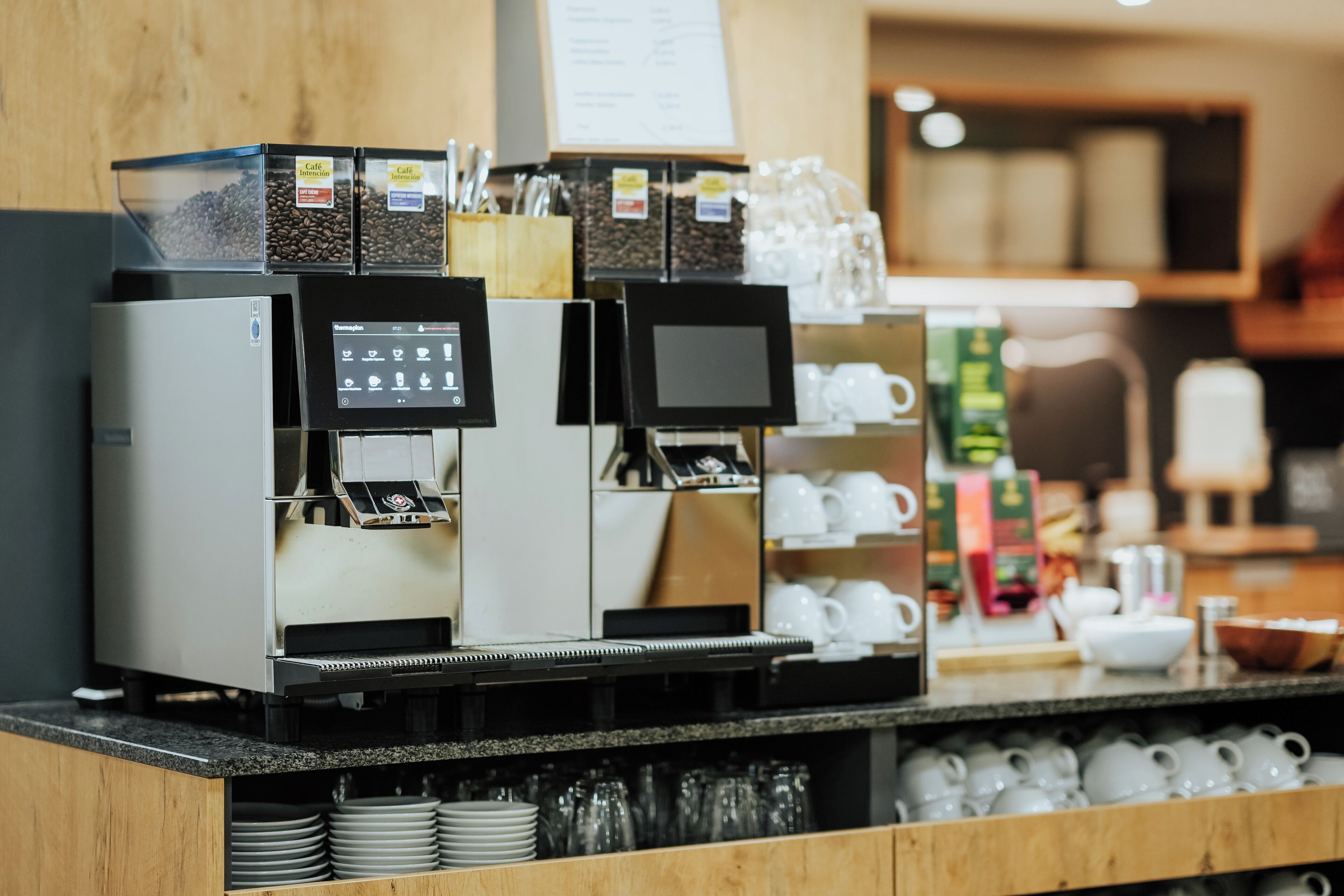A coffee machine with multiple dispensers and a digital display, coffee beans, and a variety of cups and saucers on shelves.