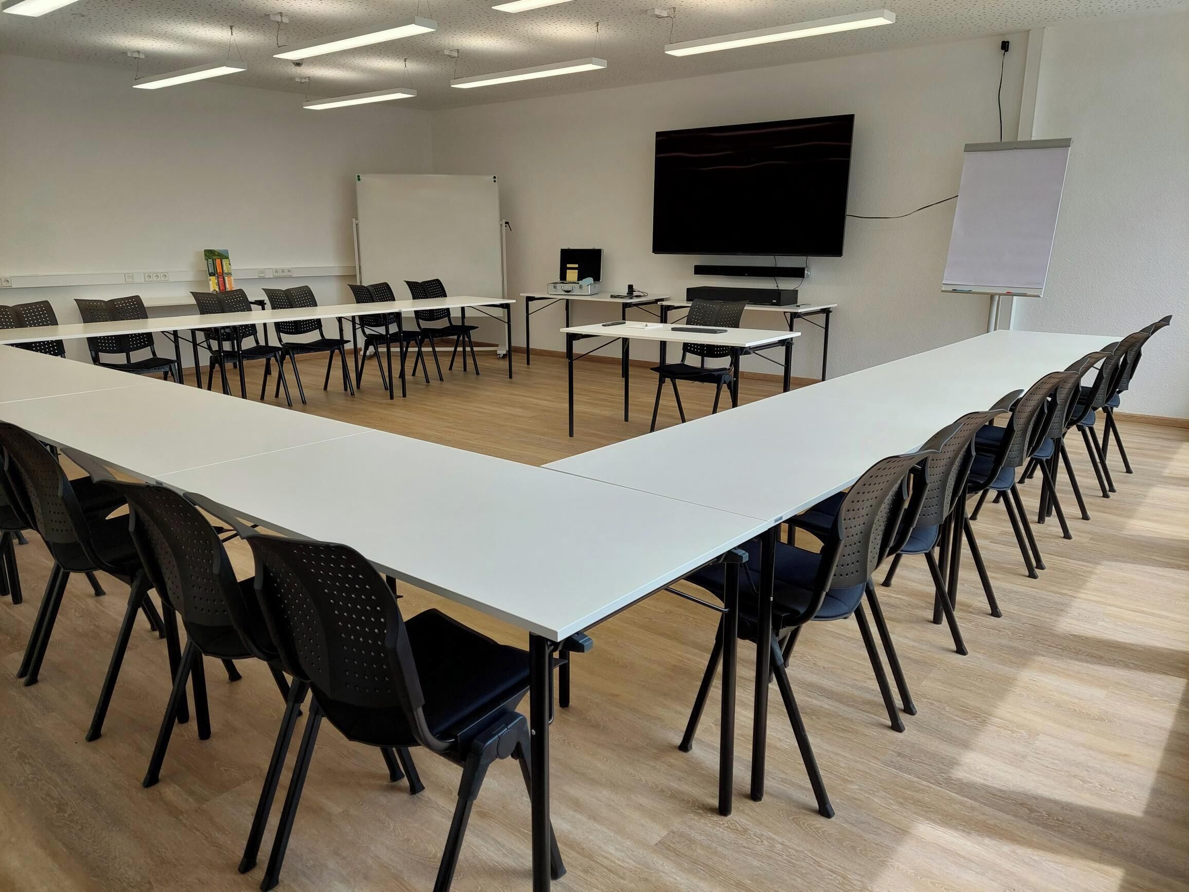 Ein Konferenzraum mit einem langen weißen Tisch, schwarzen Stühlen, einer Whiteboard-Tafel, einem Projektor und einem Fernseher an der Wand.