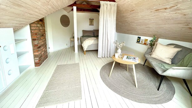 A cozy attic bedroom with a wooden ceiling, a brick wall, a bed with a white canopy, a round coffee table with a vase, and a grey sofa. - The farrier's yard (Hörby)