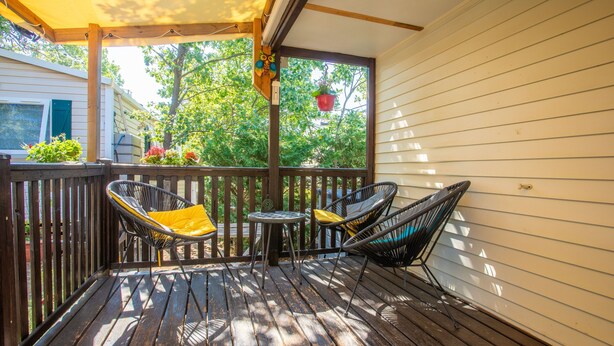 A wooden deck with two chairs and a small table, a hanging plant, and a view of trees and a house. - ‘Salamandre’ Mobile Home with Mountain Views, Shared Pool, and Air Conditioning (Sainte-Croix-du-Verdon)