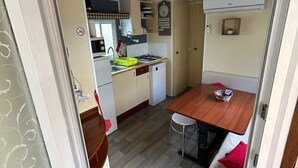 A compact kitchen with a dining area, a clock on the wall, and a small plant on the wall. - ‘Salamandre’ Mobile Home with Mountain Views, Shared Pool, and Air Conditioning (Sainte-Croix-du-Verdon)