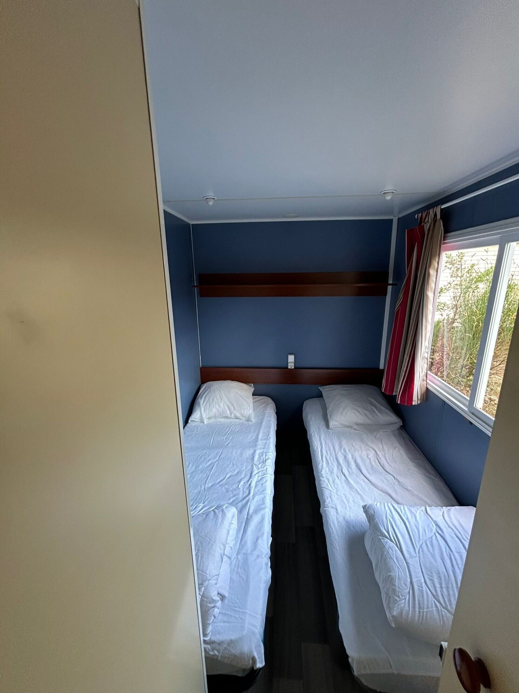 Une chambre avec deux lits simples, une tête de lit en bois, une fenêtre avec des rideaux et une porte visible.