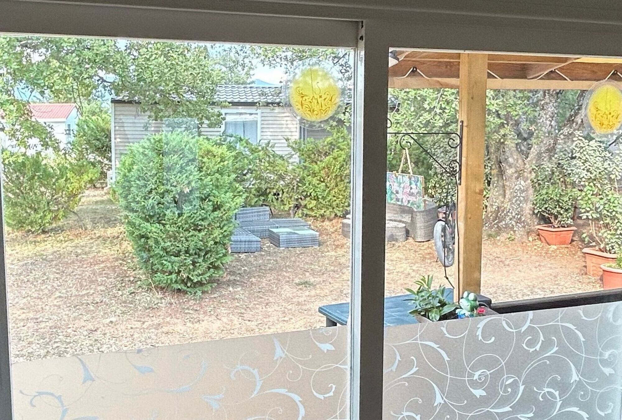 Blick durch ein Fenster auf einen Garten mit einer Holzterrasse, einer kleinen Gartenhütte und verschiedenen Kübelpflanzen.