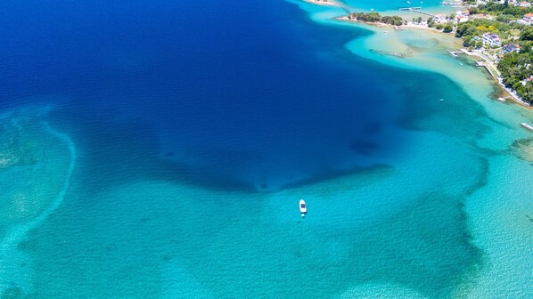 An aerial view of a coastal area with clear blue waters, a small boat, and a distant shoreline with buildings.