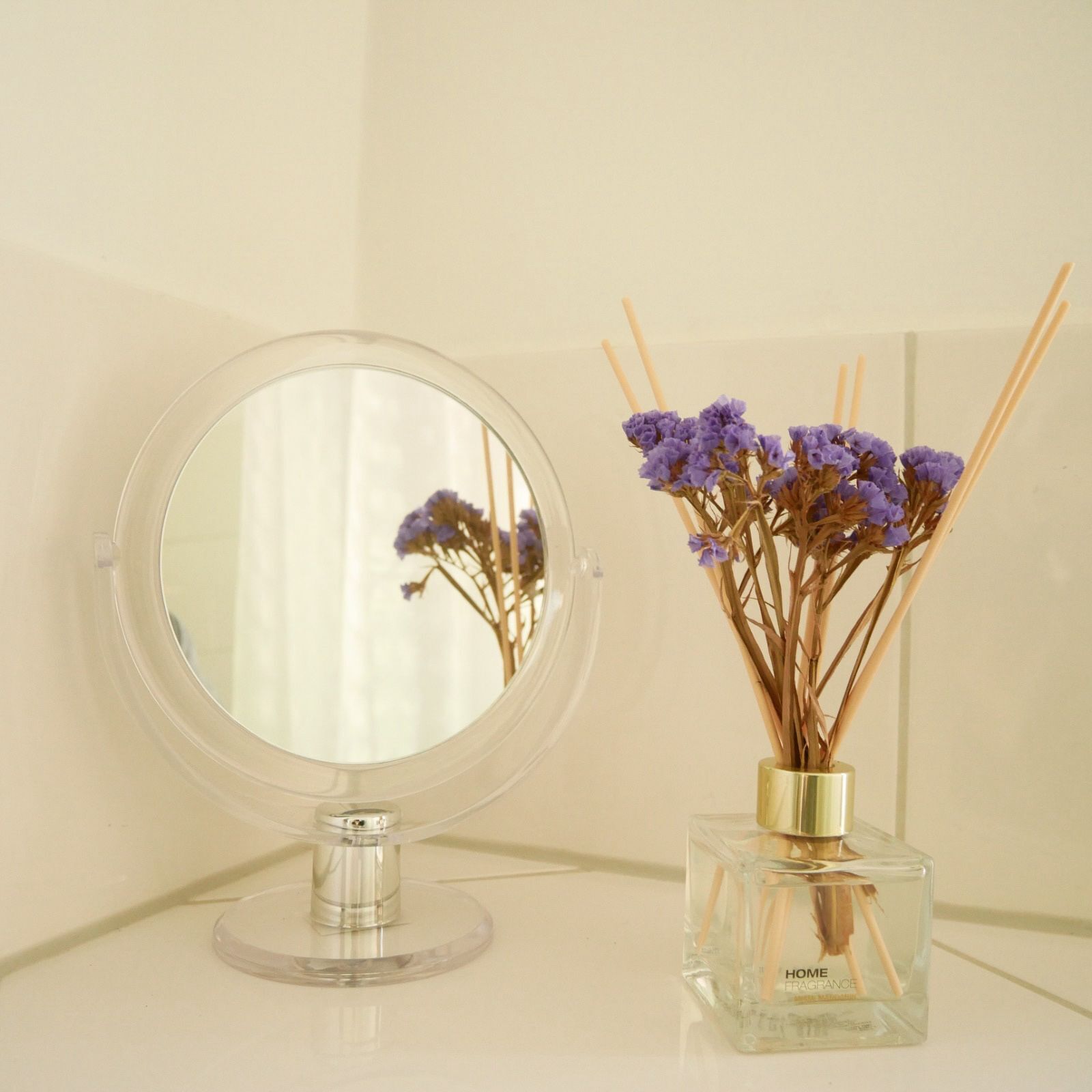 A round mirror with a clear glass base on a white surface, next to a square glass bottle with a gold cap and a bouquet of dried purple flowers.