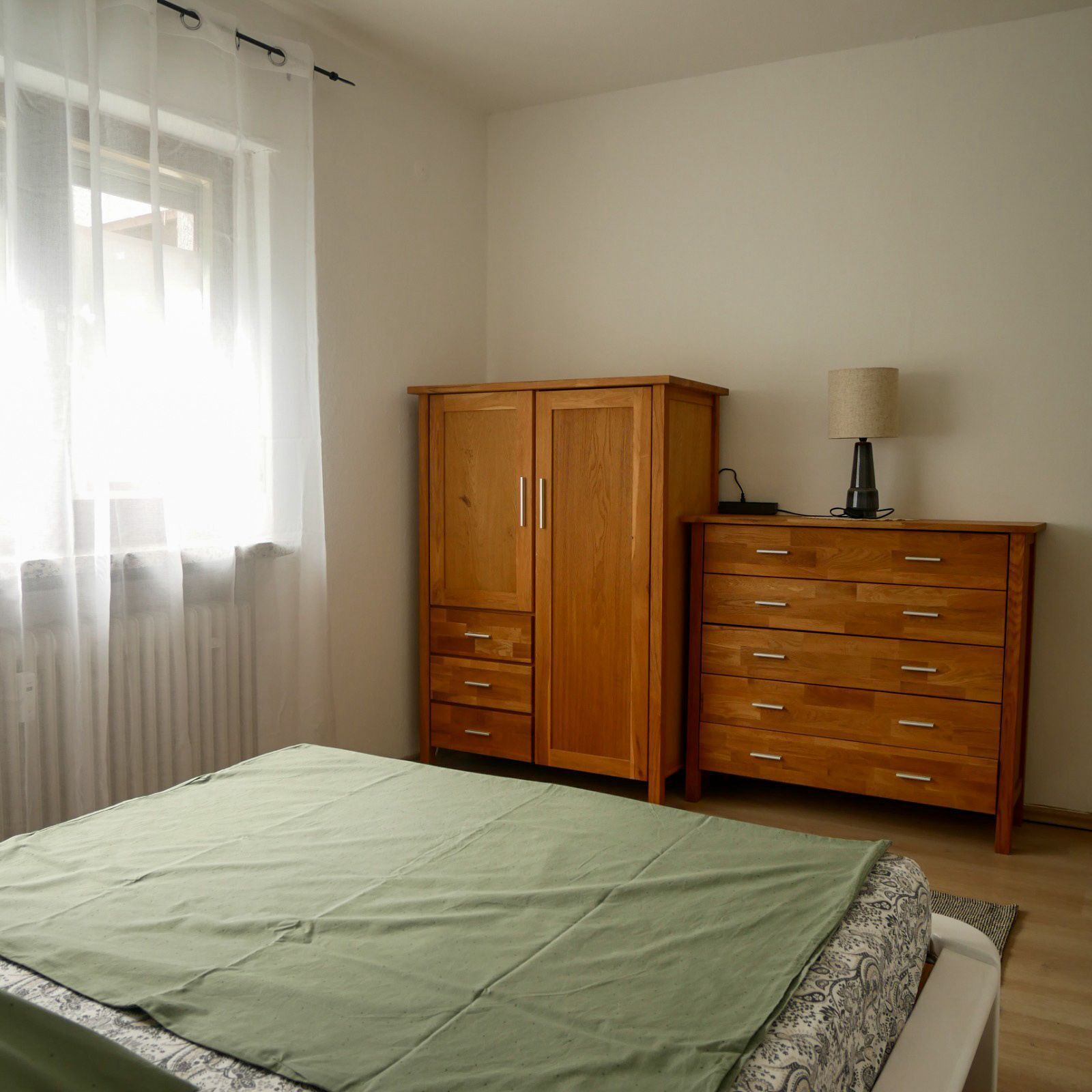 Une chambre à coucher avec un lit, une armoire en bois, une table de chevet avec une lampe et une fenêtre ornée de rideaux légers.