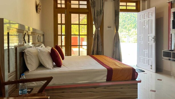 Un dormitorio con cabecera de madera, cama con ropa de cama blanca y roja, un banco de madera y un gran ventanal con cortinas.